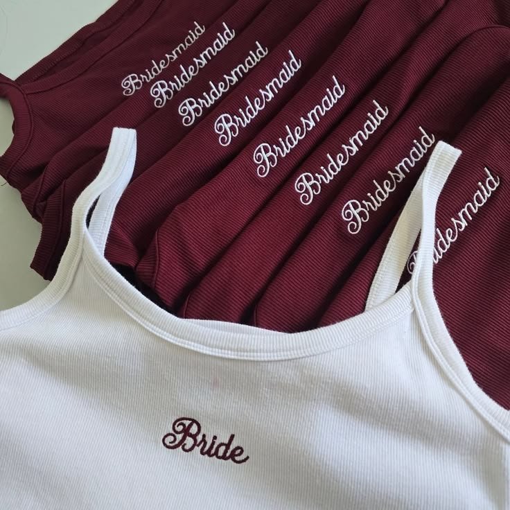 A white tank top embroidered Bride in burgundy, laid over burgundy tops embroidered Bridesmaid