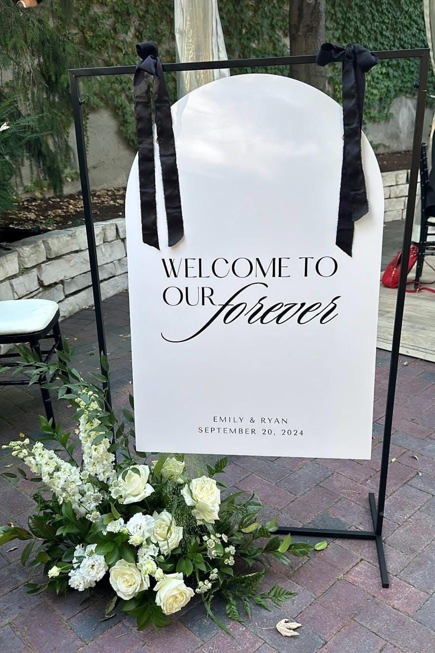 An arched white welcome sign reading Welcome to our forever, with black ribbons and white flowers at its base